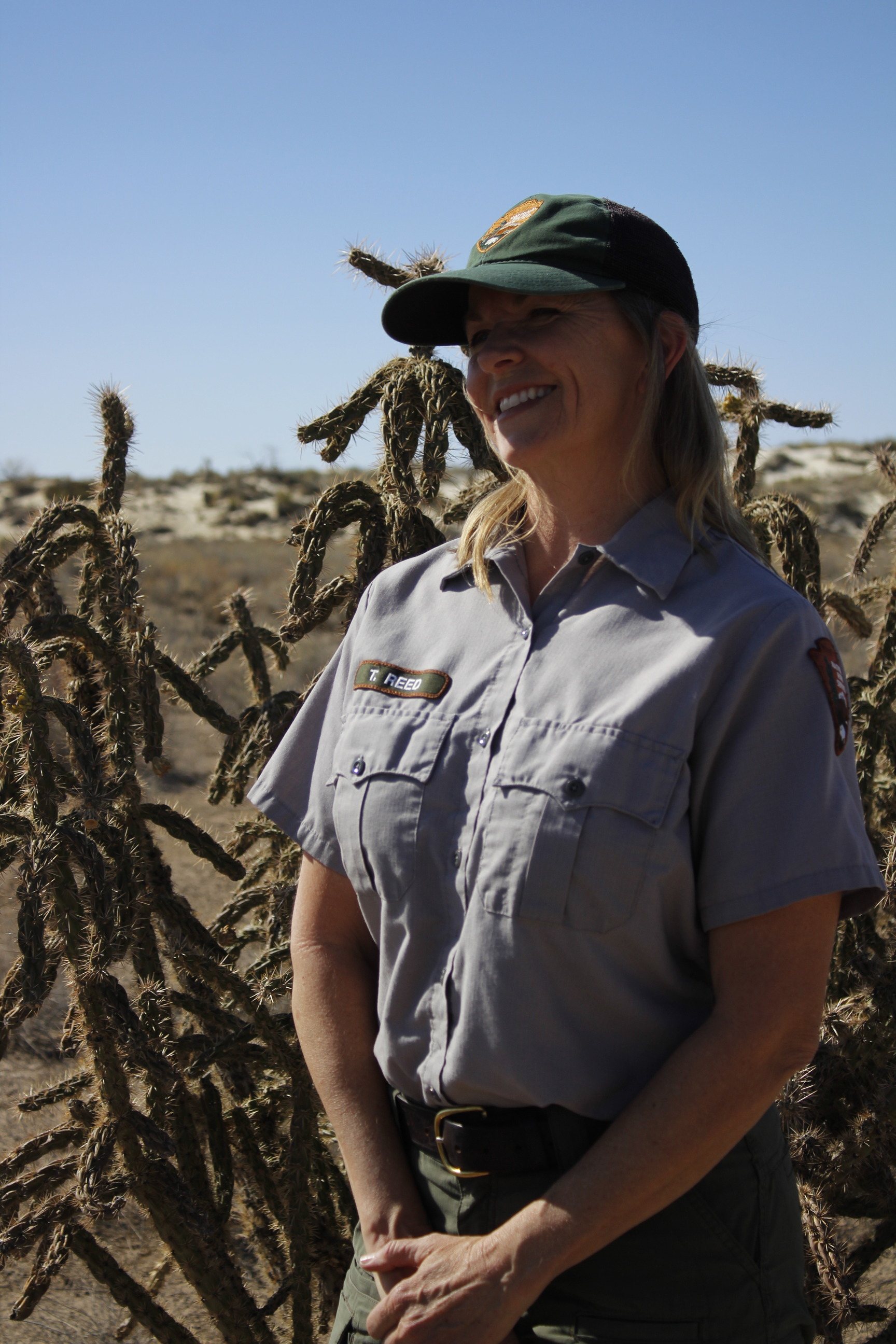 A woman in the National Park Service maintenance uniform stands for a photo. Desert cactus and shrubbery extend into the background meeting white sand dunes under a clear light blue sky. 