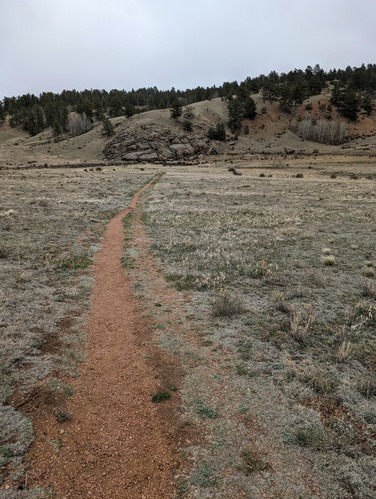 A granite trail crosses a grass filled valley and approaches a hillside with large rock feature.