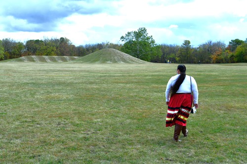 A lady wearing a white blouse and colorful red skirt with alternative horizontal colorful stripes walks in a green grass field with large earthen, green grass-covered mounds in the distance.