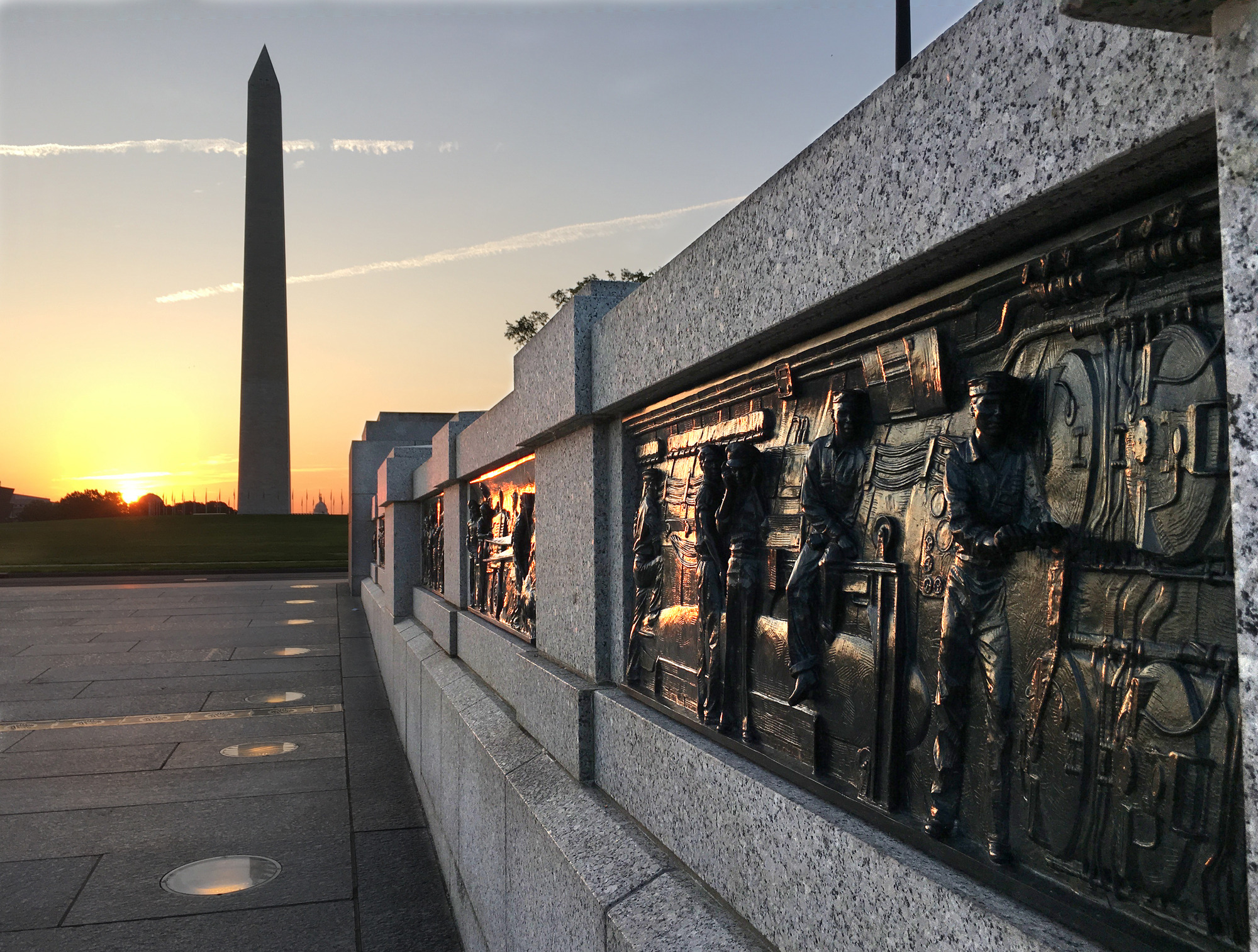 Dark, bronze, rectangular panels set into a light stone wall that stretches from the bottom right to the center of the photo, in the direction of the Washington Monument, a tall obelisk in the distance. The sun is on the horizon to the left of the monument. 