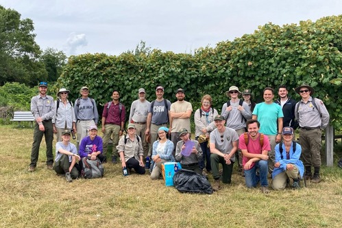 A group of people of varying ages standing and kneeling together for a group photo. Some of the people have NPS uniforms on. Behind them, the Grape Island grape arbor is covered in grape vines 