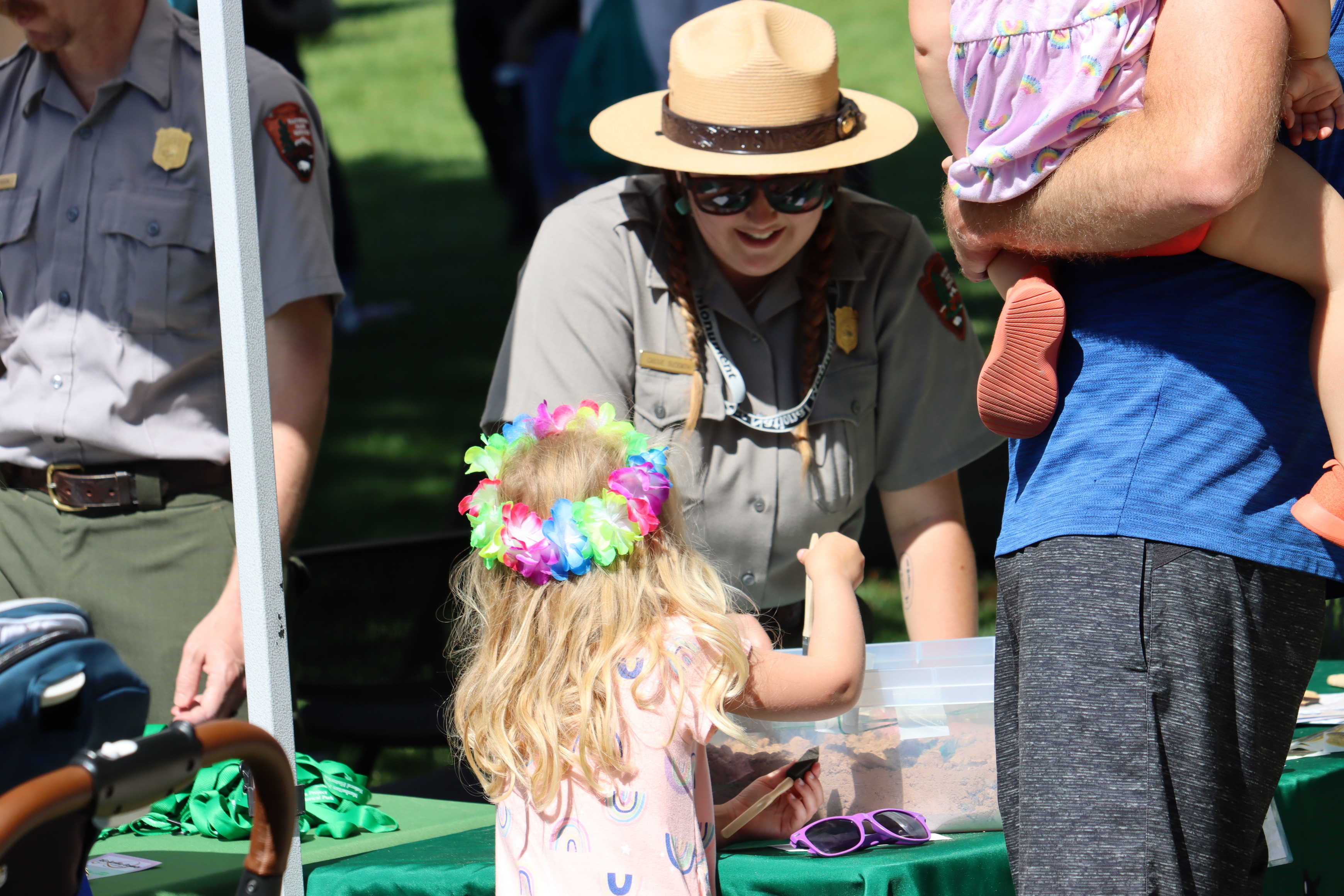 Uniformed park ranger smiles and looks down towards a young blonde girl with a flower crown as she digs in a box of sand on a table. Another ranger in the background and an adult holding a smaller child in his arms at a community tabling event.