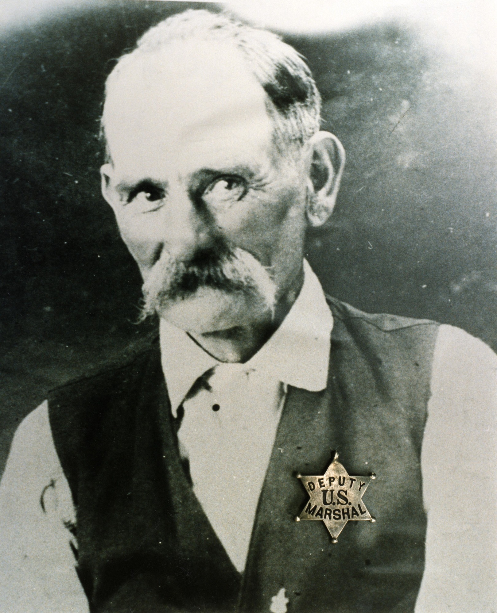 Older white man with thinning hair, and a thick mustache wearing a white shirt, dark vest, and gold star imprinted with text Deputy US Marshal.
