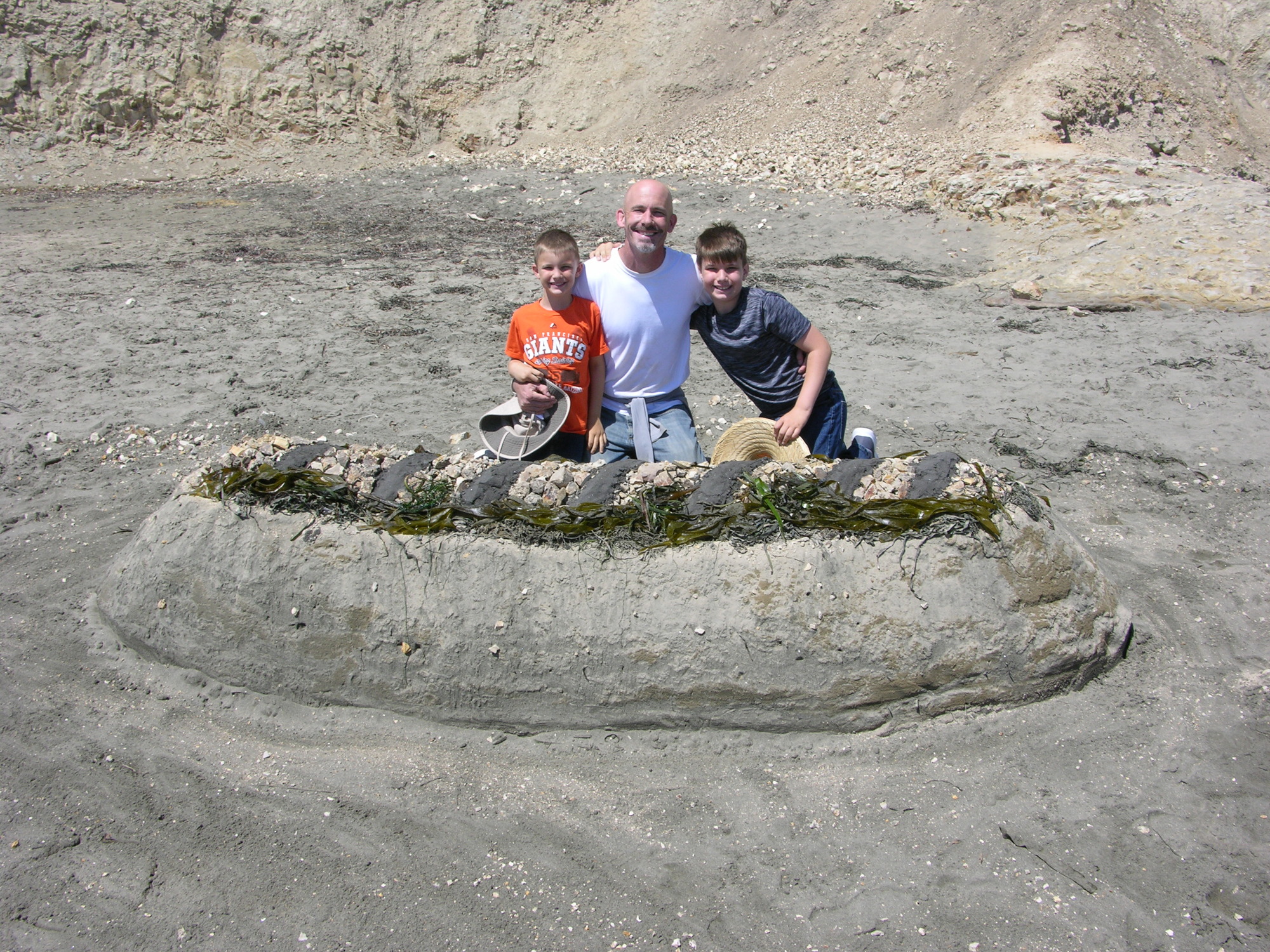 A man and two boys behind a sand sculpture of a large hotdog.
