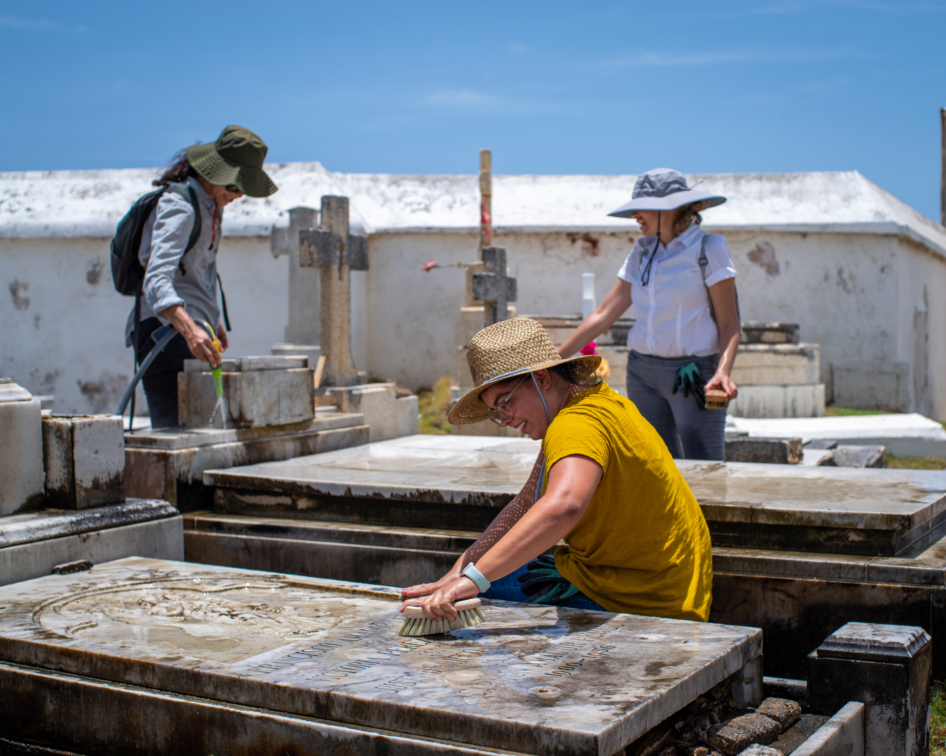Woman scrubbing top of tomb with two women rinsing another tomb in the background