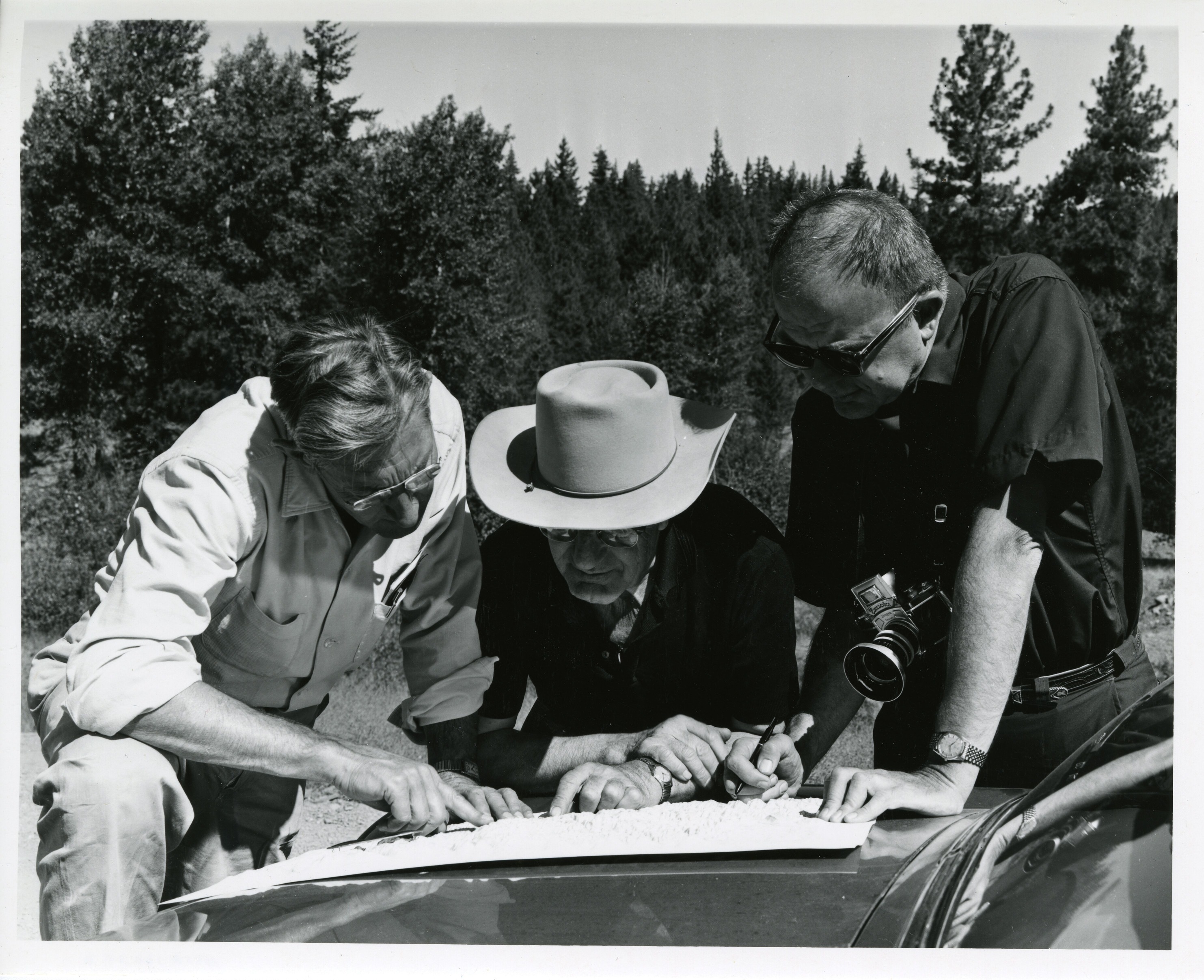 Three men looking over a map layed out on the hood of a car. Coniferous tree line in the background.