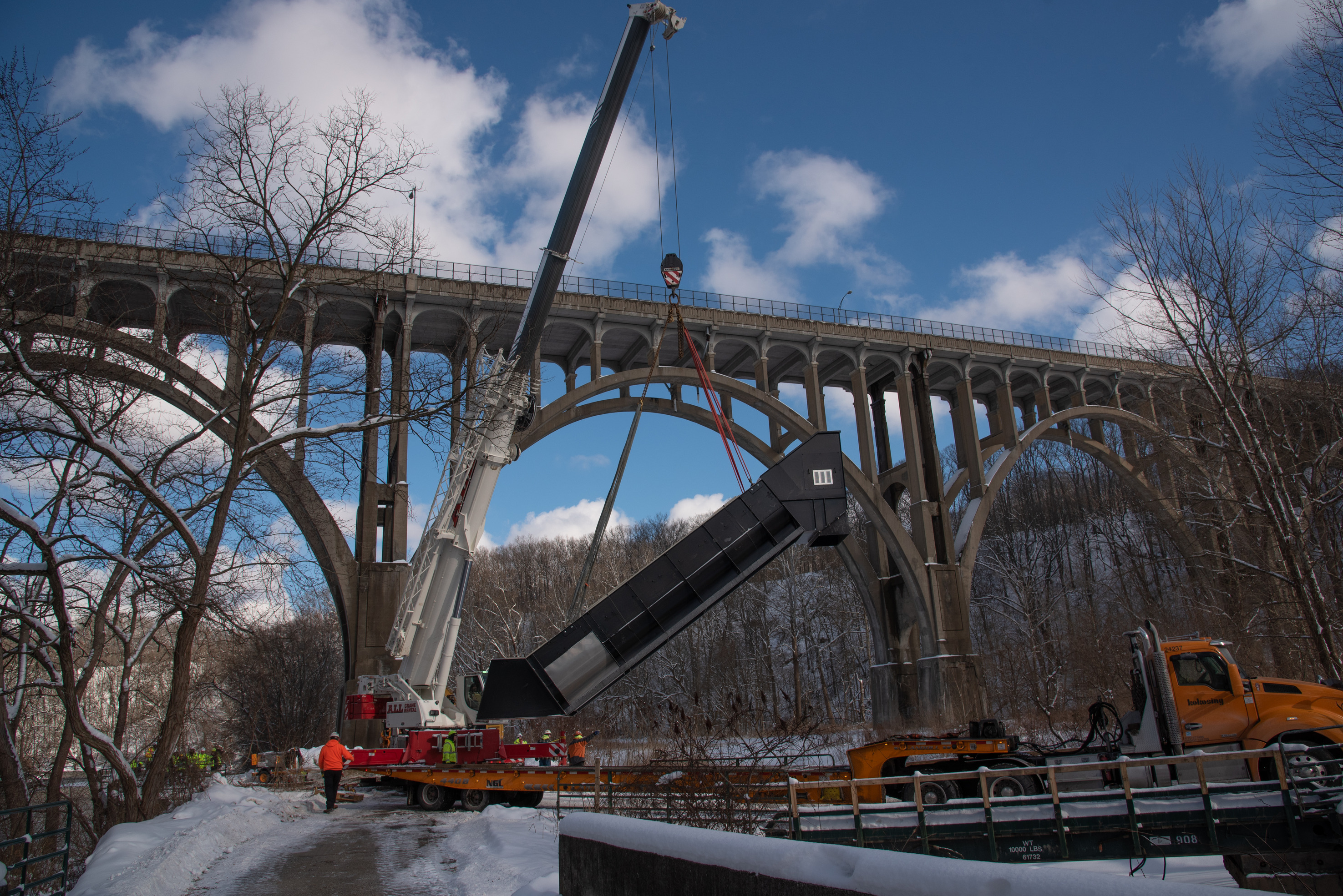 The brown, metal pump is a long boxy tube at a 45 degree angle. It is suspended over an orange truck. Workers cluster nearby.