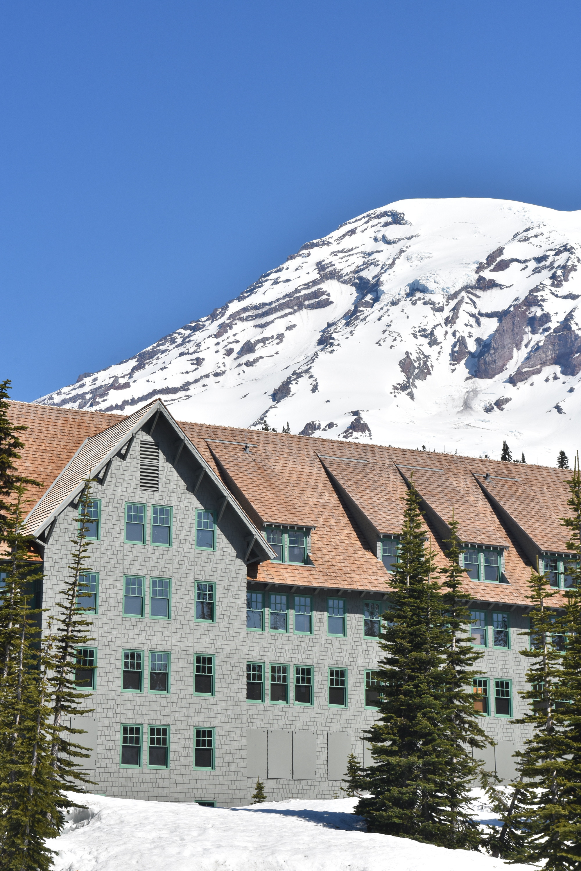 A four-story wood building with grey shingled sides and an angled roof in front of a glaciated mountain. 
