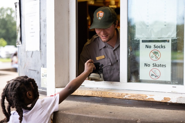 A young boy holds out his sneaker to show his shoe size to smiling National Park Service Park Guide Hunter Baldridge at the skate rental booth at Anacostia Park Skating Pavilion in Washington, D.C., on June 28, 2025.