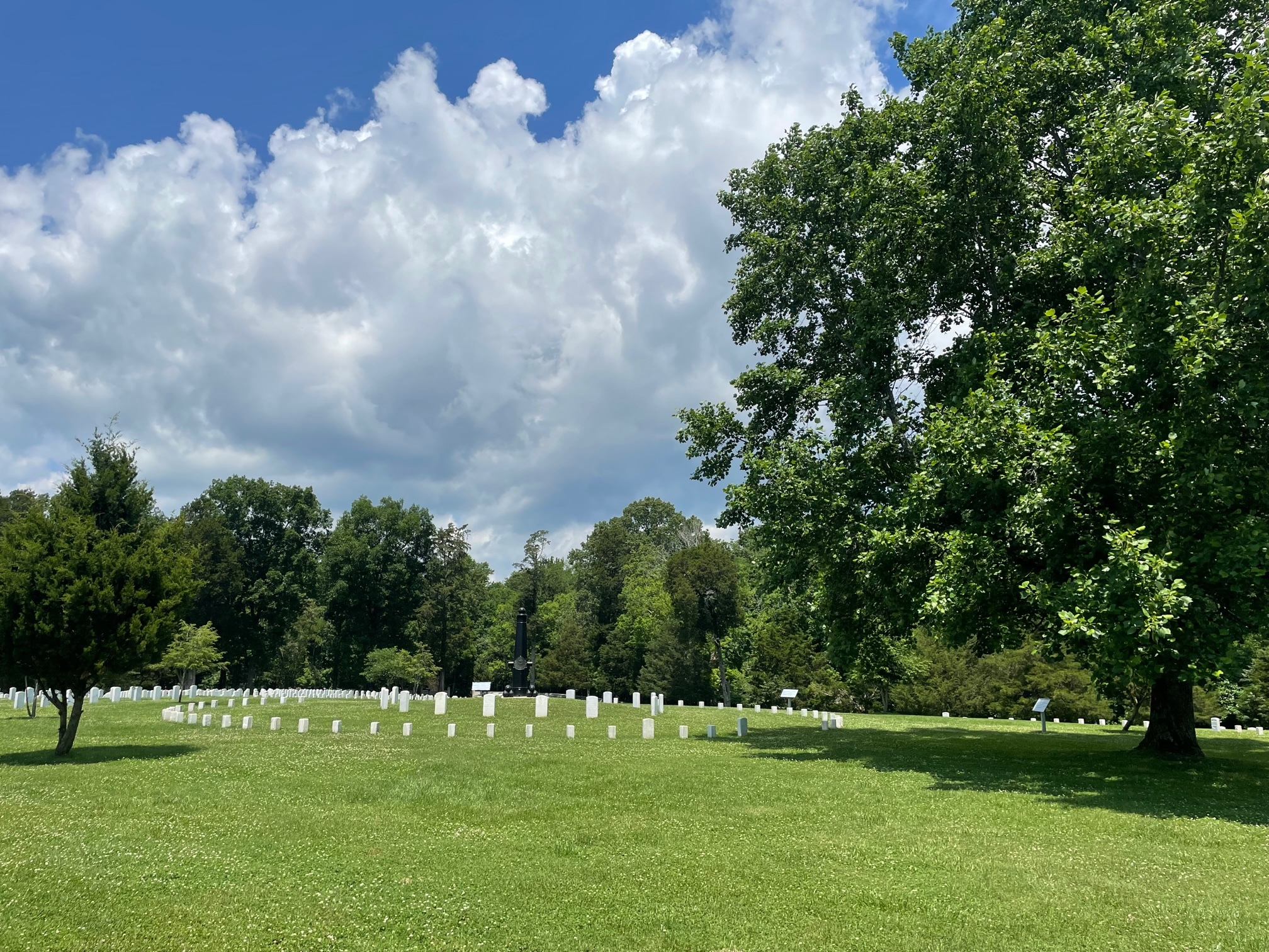 Uniform white grave markers in a national cemetery form a circle around a monument, which is a black upright cannon. Leafy trees shade the turf. 