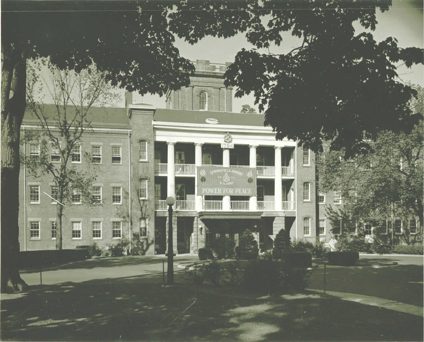 Black and white photo of part of the front of a large brick building with a second and third story balcony and a tower in the back. An insignia and the year “1794” hangs above the middle of the third story balcony. A banner hangs from the third story balcony and reads “Springfield Amory, U.S. Army, Power for Peace.” Trees and bushes grow around the building.