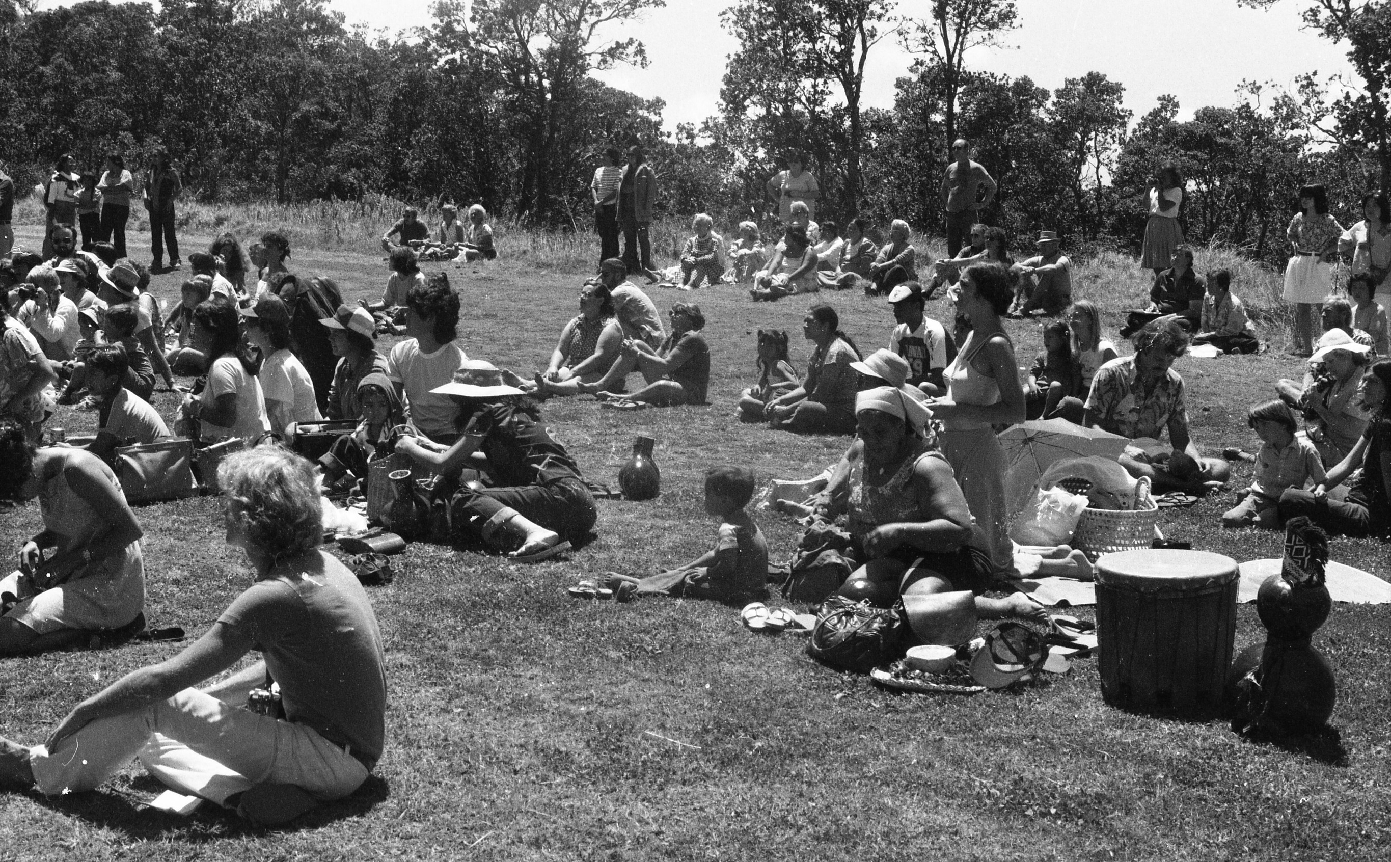 A black and white image of a large crowd of people sitting on the grass. The people are spread out evenly across the image. Some are sitting down on the grass while others are standing behind the people sitting down. Everyone is looking towards the left side of the image. Behind the crowd there is a dense wooded area.