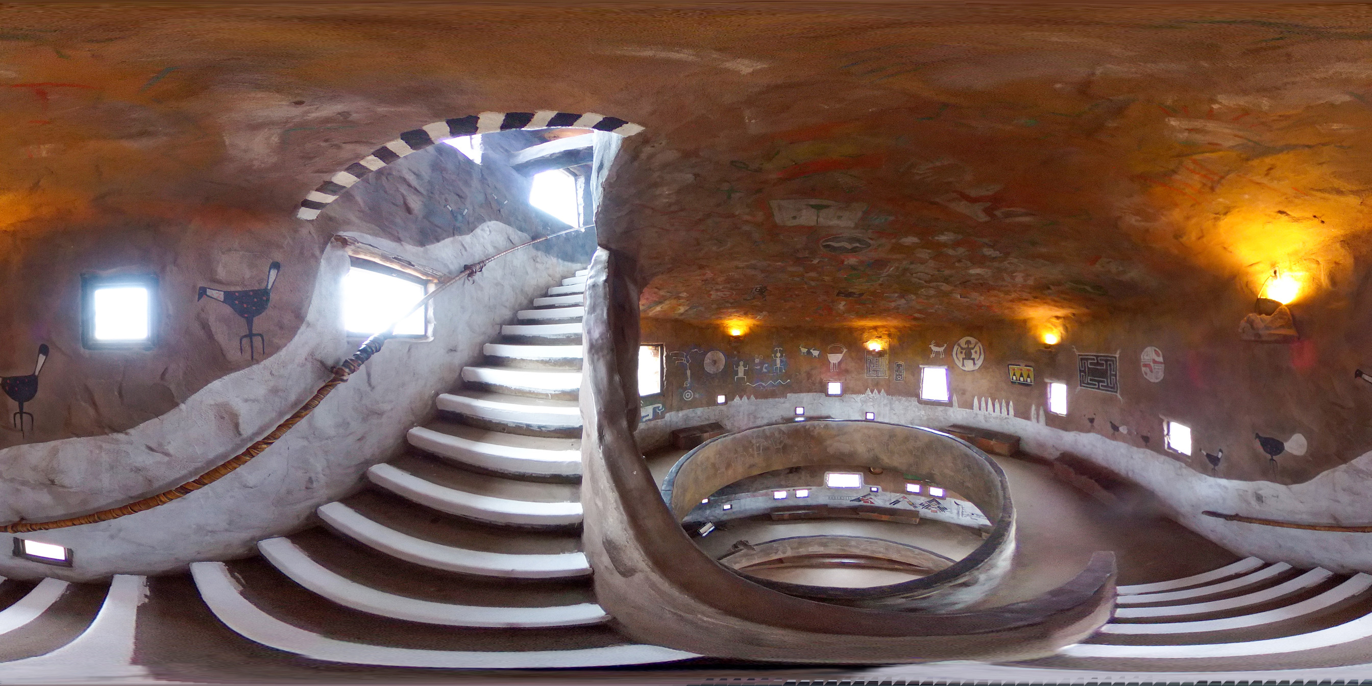 Within a circular tower, from a spiraling stairway,  with a ceiling decorated with Ancestral Puebloan designs. Across the room,  murals wrap around the circumference of the room. Through a circular parapet in the center, two additional levels are visible below.
