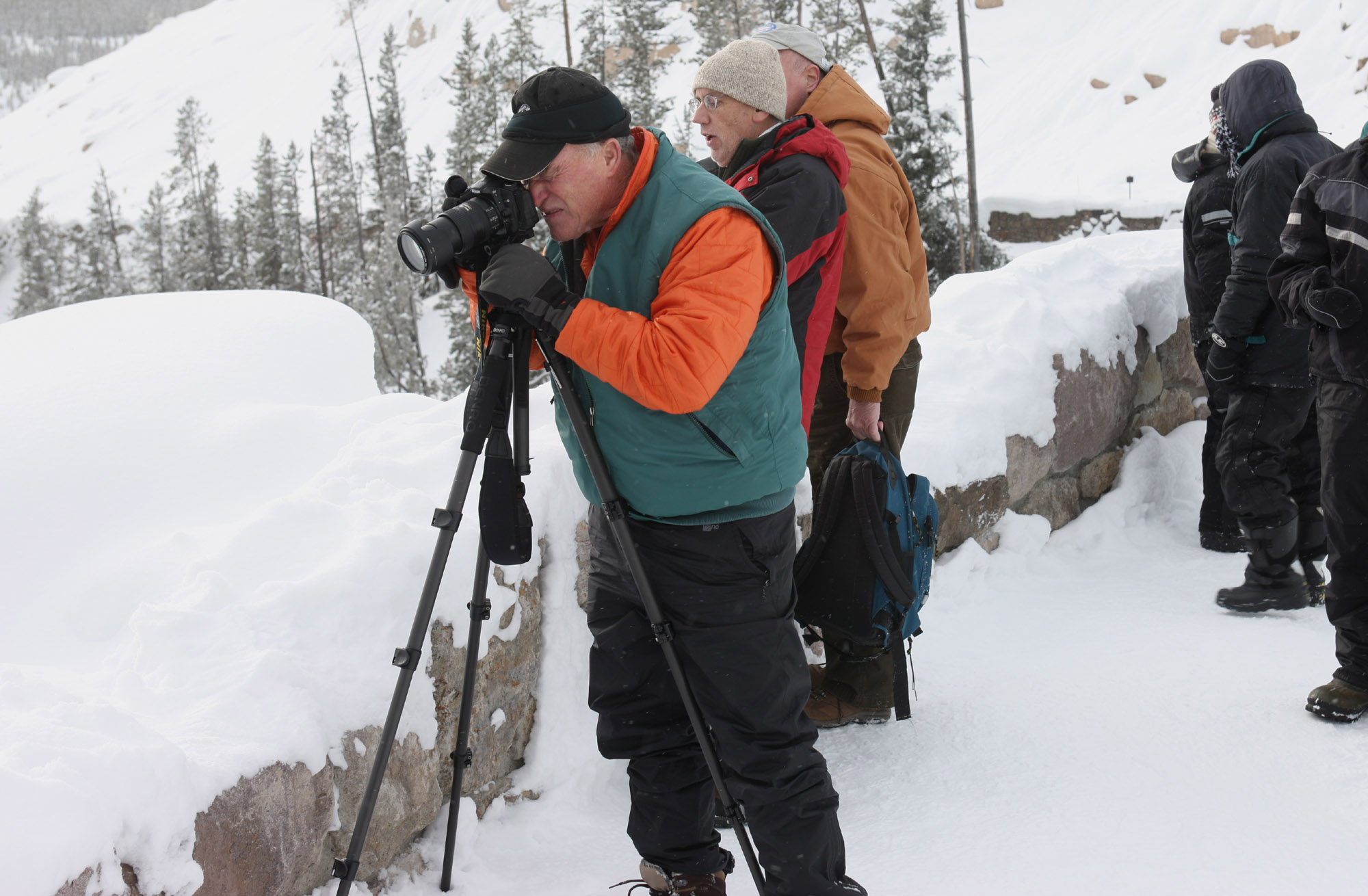 A man is pointing a camera on a tripod to the left and he is looking through the view finder in winter with snow on the ground.