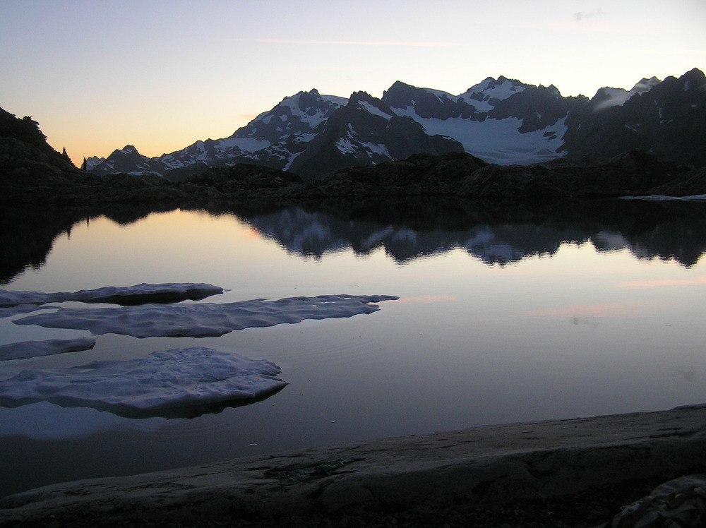 A calm mountain lake at sunset, with patches of snow floating on the surface near shore.