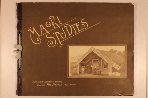 Front cover of late 19th century photo album with the title 'Maori Studies'. Bottom left corner with the words 'Permanent Carbonette Photos. Copyright. Thos. Pringle. Wellington.' Bottom right corner photograph of Maori home with three women in front.