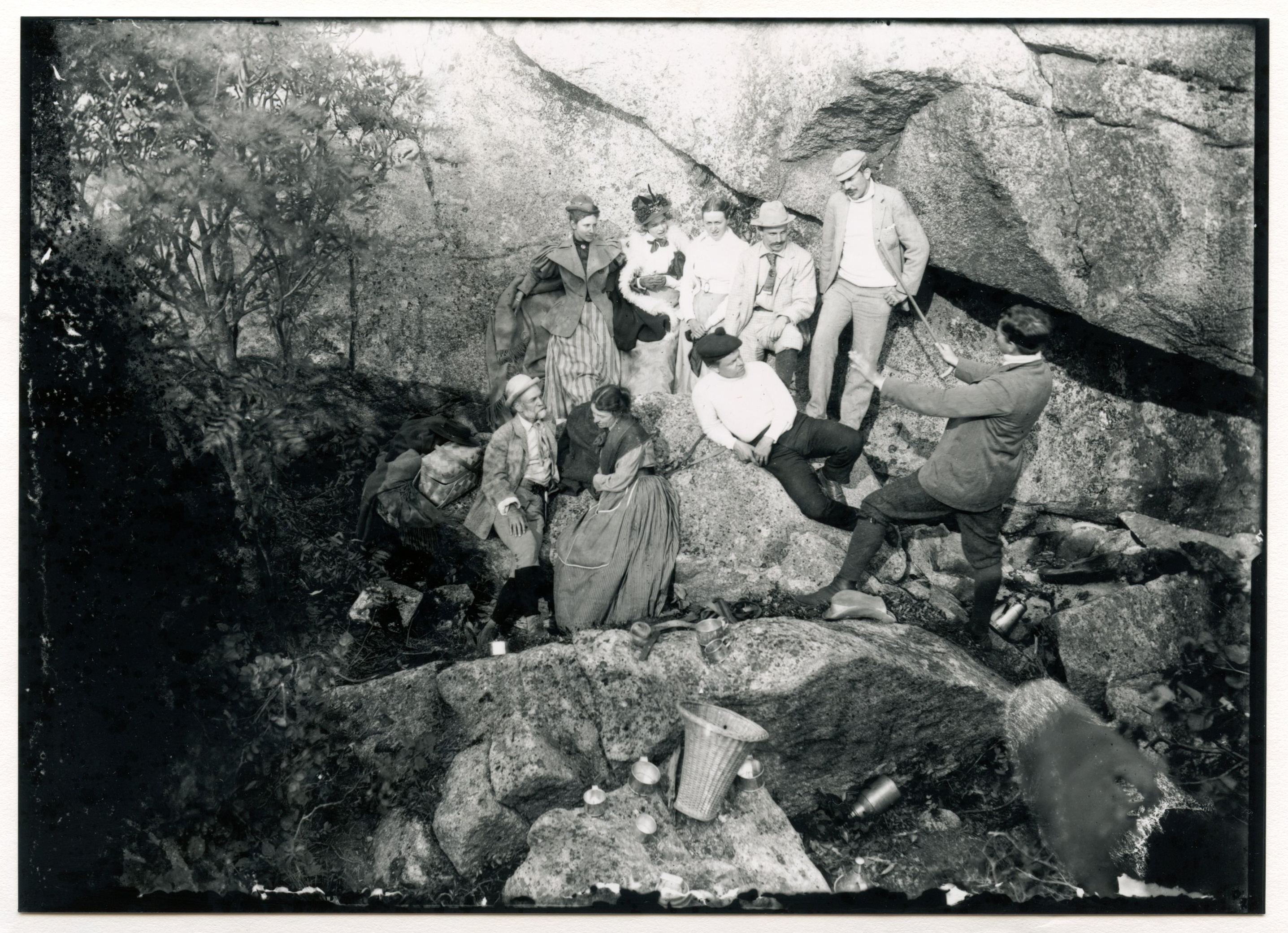 White men and women relax next to a large rock feature. They have wicker baskets and metal containers. One man stands at the front of the group facing everyone and holds out his arms, a cane in his right hand. Trees at left.