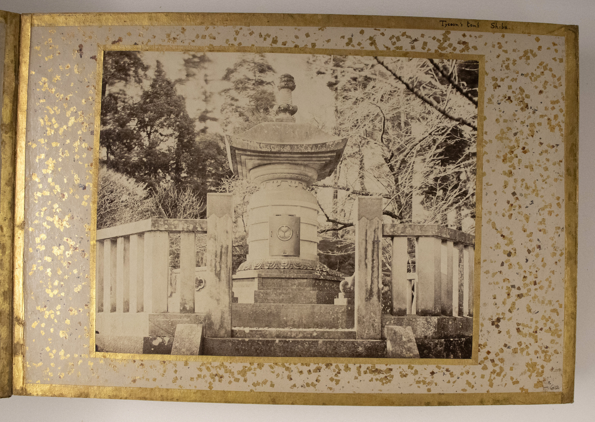 Sepia photograph representing a stone tomb elevated on a platform surrounded by a fence and trees.  The photograph is placed in the center of an album decorated with a golden leaf frame, dots and has inscriptions.