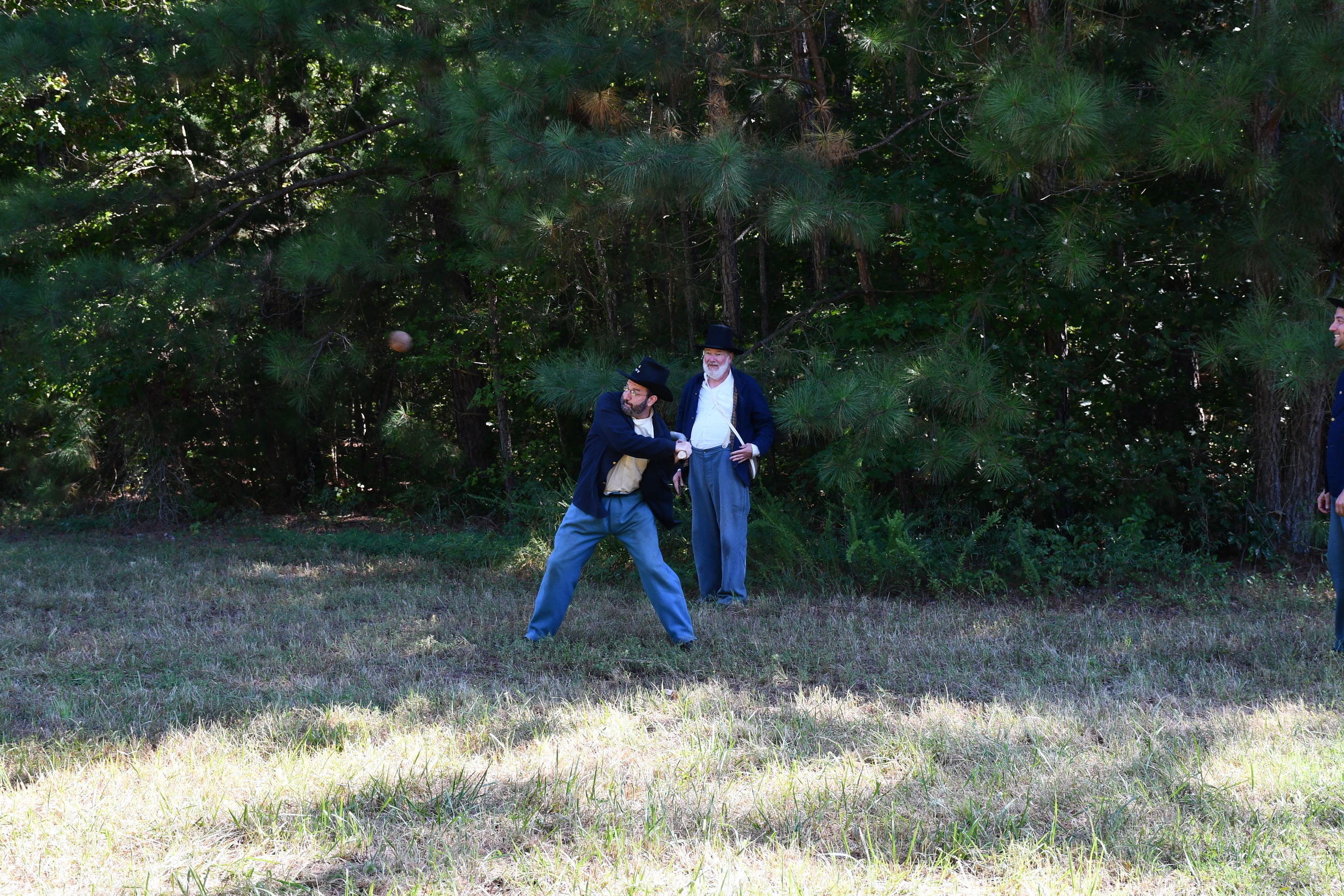 A man wearing light blue trousers, white shirt, dark blue jacket, and a dark blue Hardee hat prepares to swing a branch at a large leather ball.