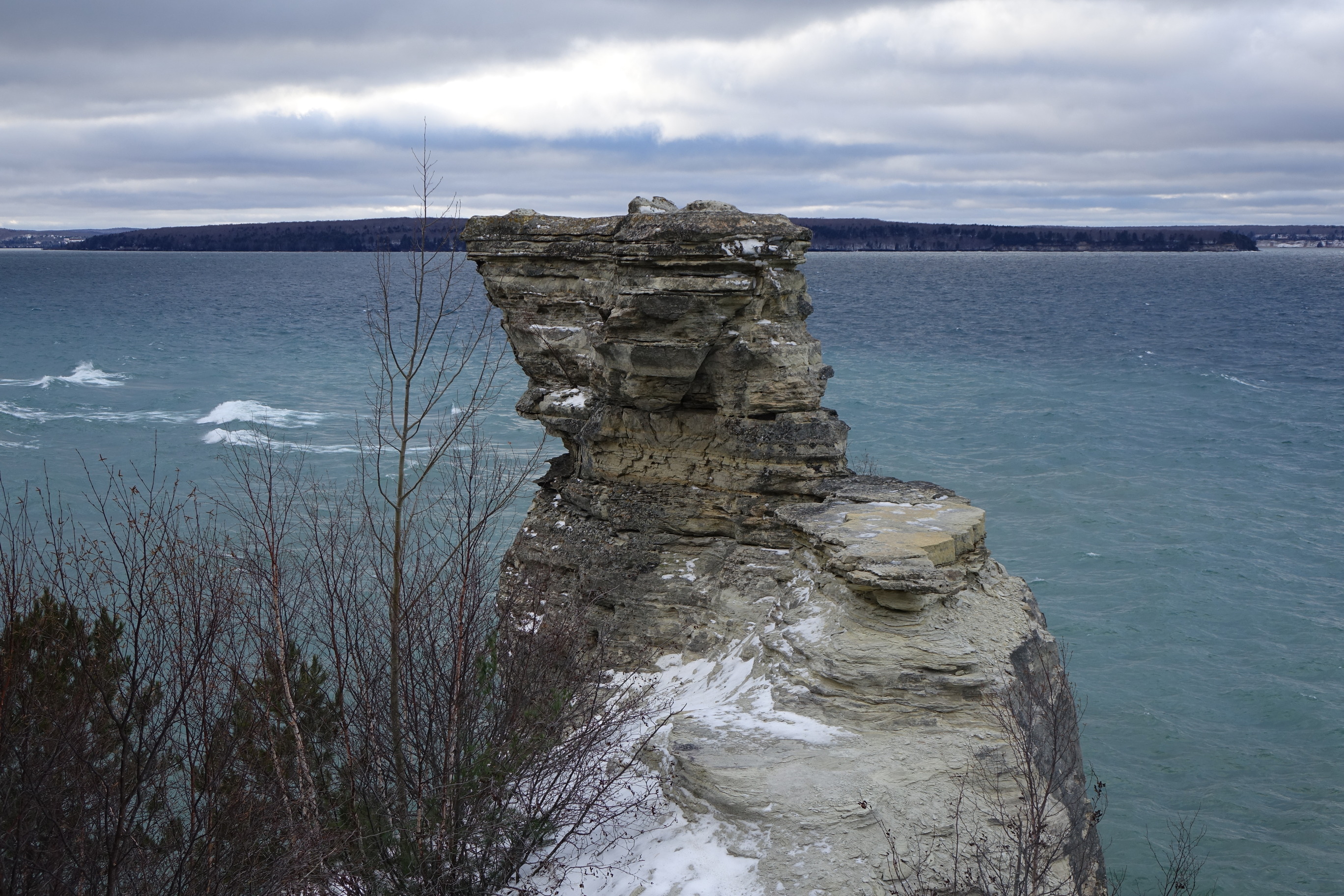 The top of the rock formation looks like a castle turret. Light snow is on the ground. Lake Superior waves are in the distance.