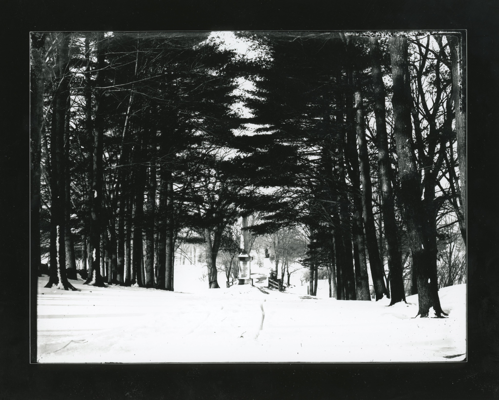 View from a snow covered path leading onto a bridge. Trees line the road. Obelisk statue before the bridge. Statue of a man past the bridge in the far distance.