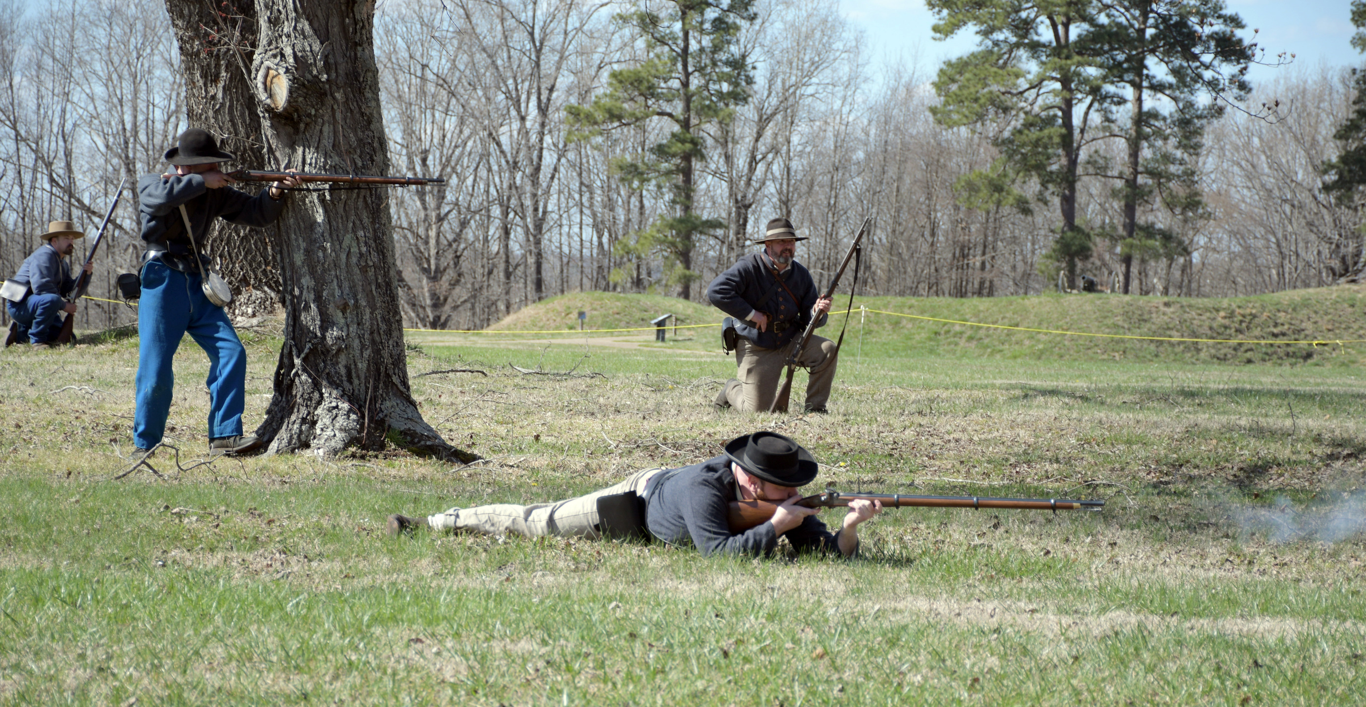 Four men wearing Confederate uniforms. Two in the background are knelling, one is standing behind a tree preparing to fire his rifle the 4th man is in a prone position having just fired his rifle as the smoke can be seen.