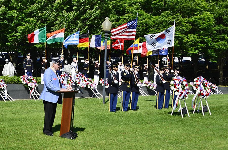 A veteran speaks at a lectern with a colorguard displaying various nations' flags.