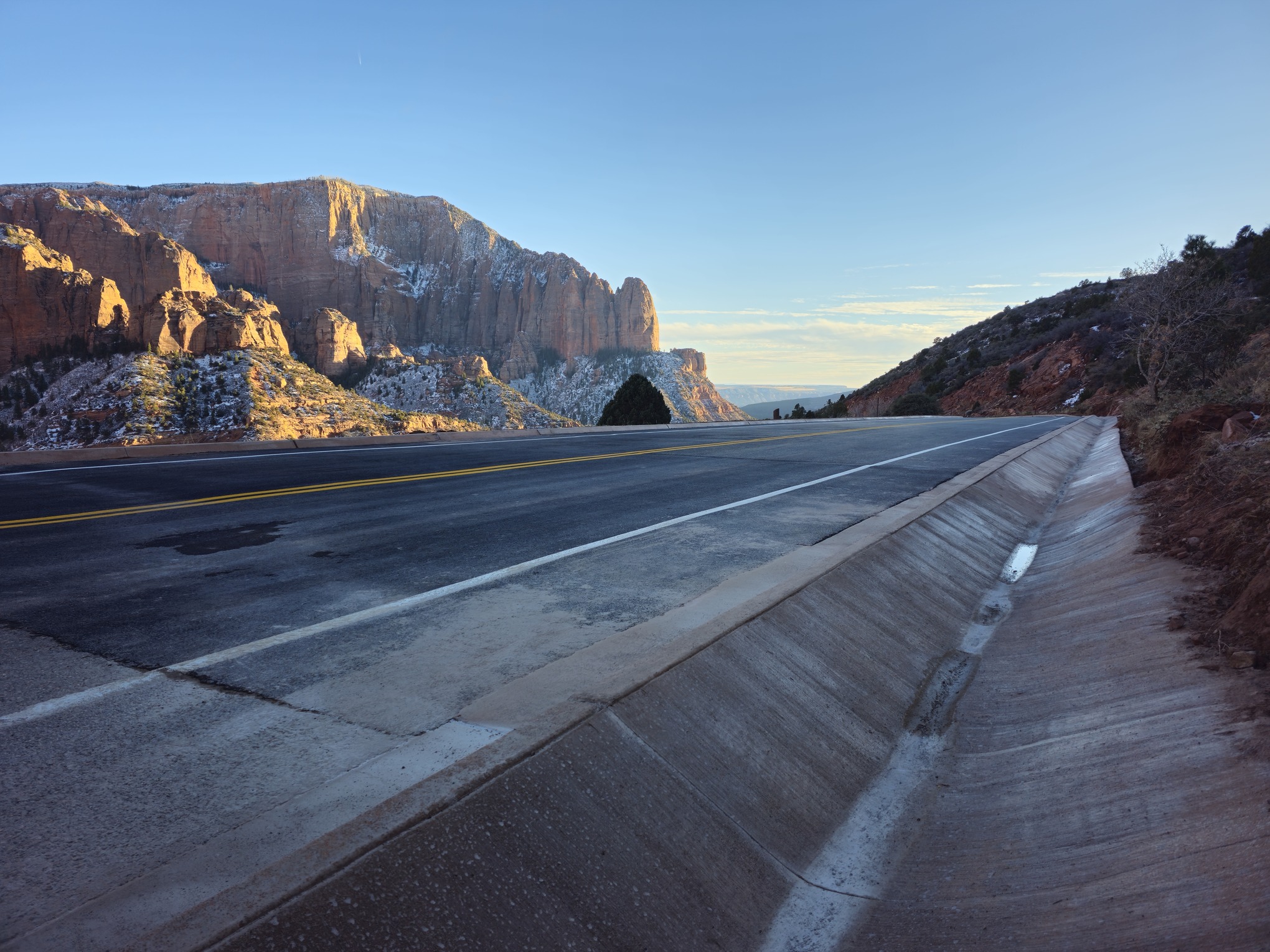 Paved drain next to a newly paved asphalt road with red rocks and snow in the distance and blue sky overhead.