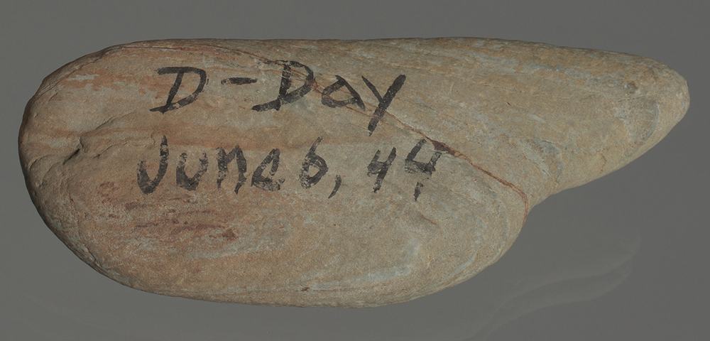 An oval light brown rock has “D-Day June 6, 44” written on it in black ink.