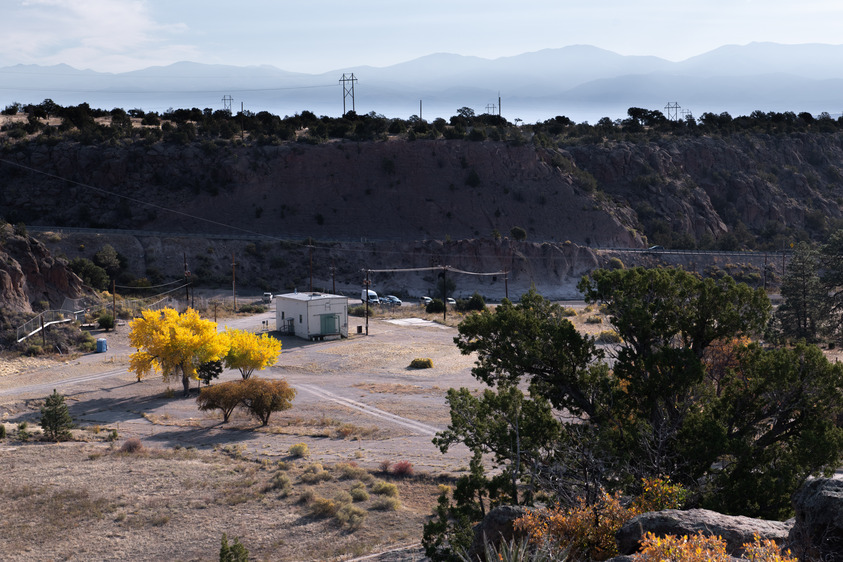 A white square building stands next to yellow cottonwood trees in the flat bottom of a dry valley, the valley walls covered in green trees. Behind, mountain ridges in hazy skies.