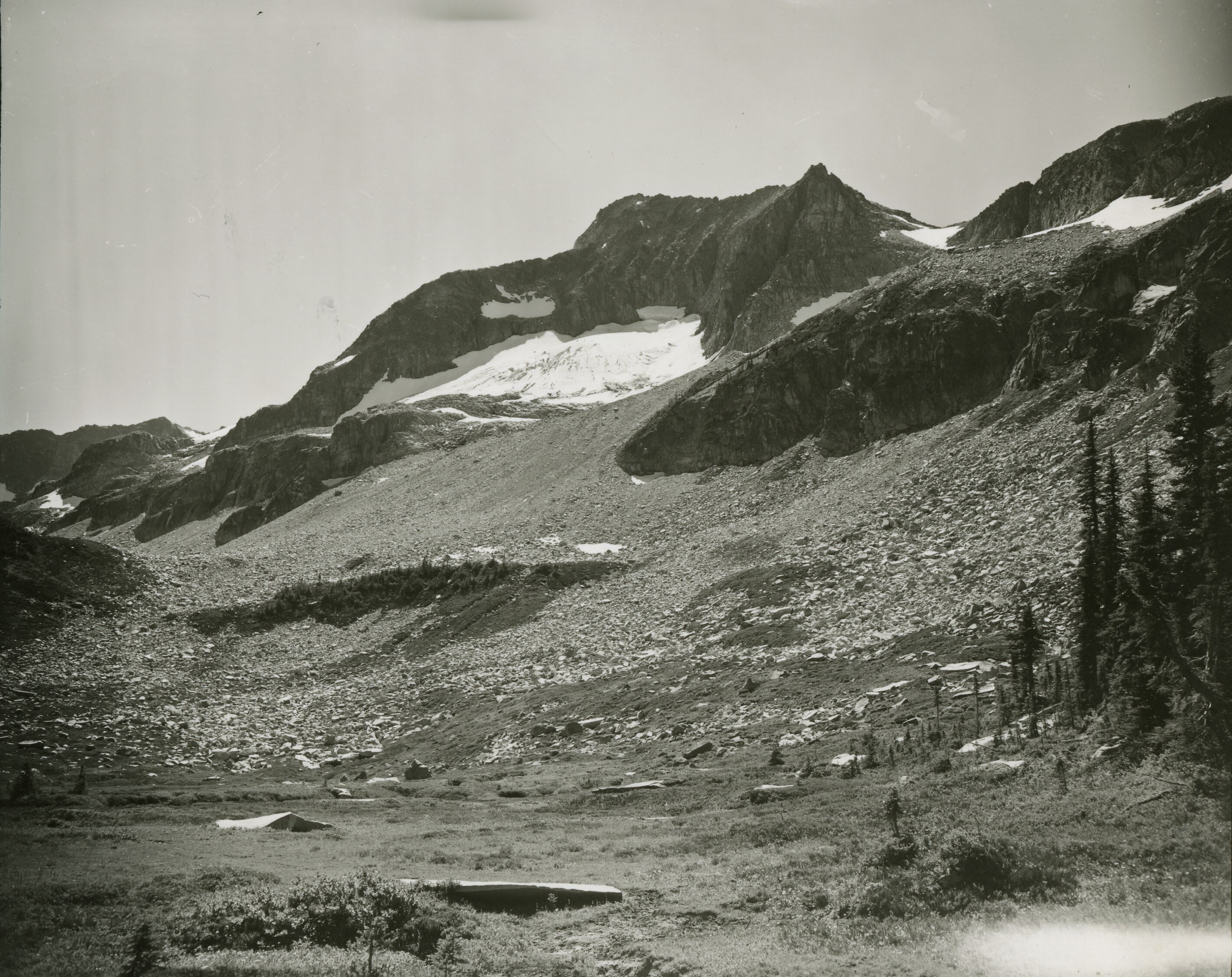 A sloping hill covered in rocks and boulders. A rocky mountain peak with snow in the background.