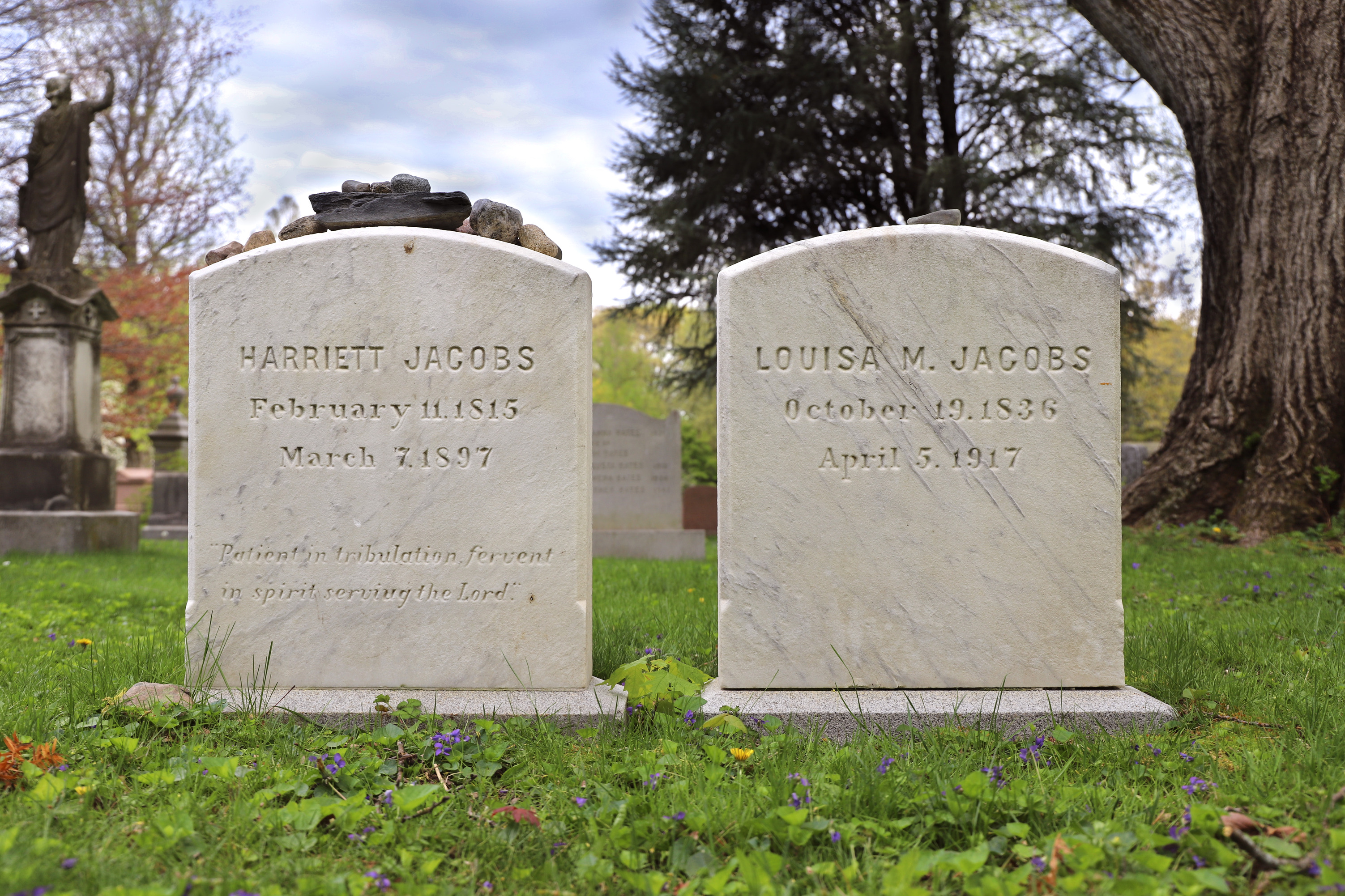Two light stoned grave stones sit next to each other. The left stone is inscribed "Harriet Jacobs, February 11, 1815, March 7, 1897, 'Patient in tribulation, fervent in spirit serving the Lord.'" Small rocks sit on top of it. The right headstone reads "Louisa M. Jacobs, October 19, 1836, April 5, 1917." Dandelions and small purple flowers pop out of the grass surrounding the headstones.