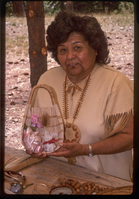 A woman in a Native American attire holds a woven basket with items inside.