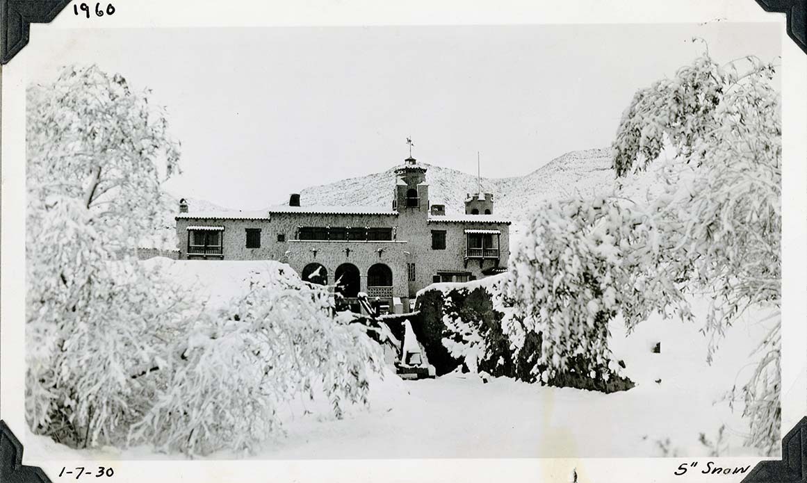 This is an historic black and white photograph from the Scotty's Castle Historic Photograph Collection, Death Valley National Park of snow covered branches to large Spanish style building with arched front porch. Central foreground white. Snow covered hill behind. Inscriptions in black ink along upper and lower border.