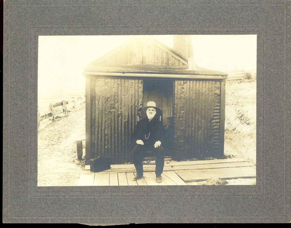 Albumen card-mounted photograph of John Burkman seated in front of his house.