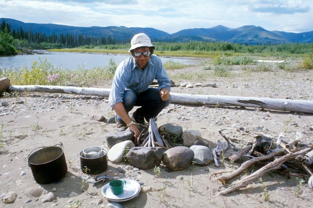 man building a fire in a rock ring on a sandy beach, near a river