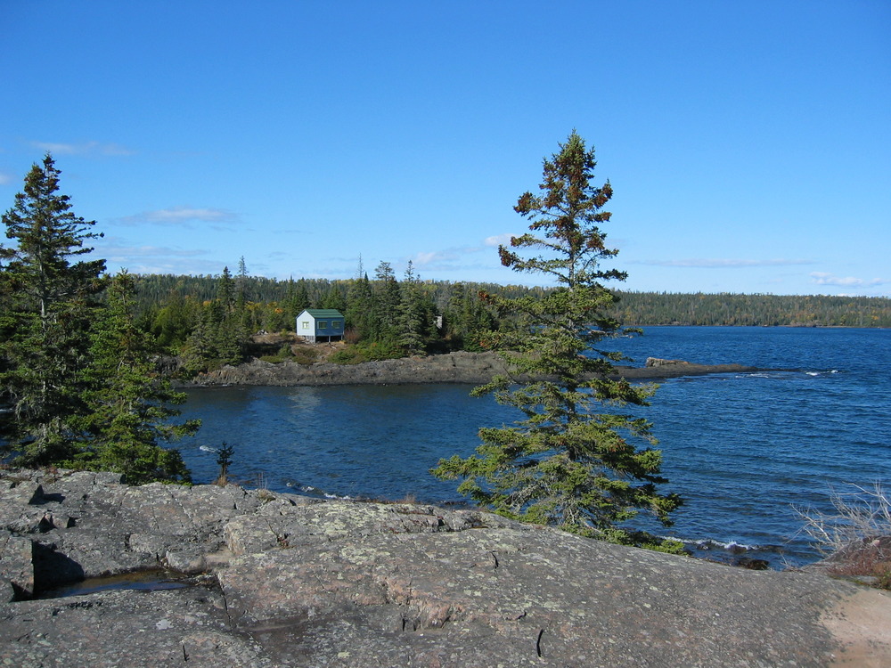 View of cabin perched on shoreline with rocky foreground.