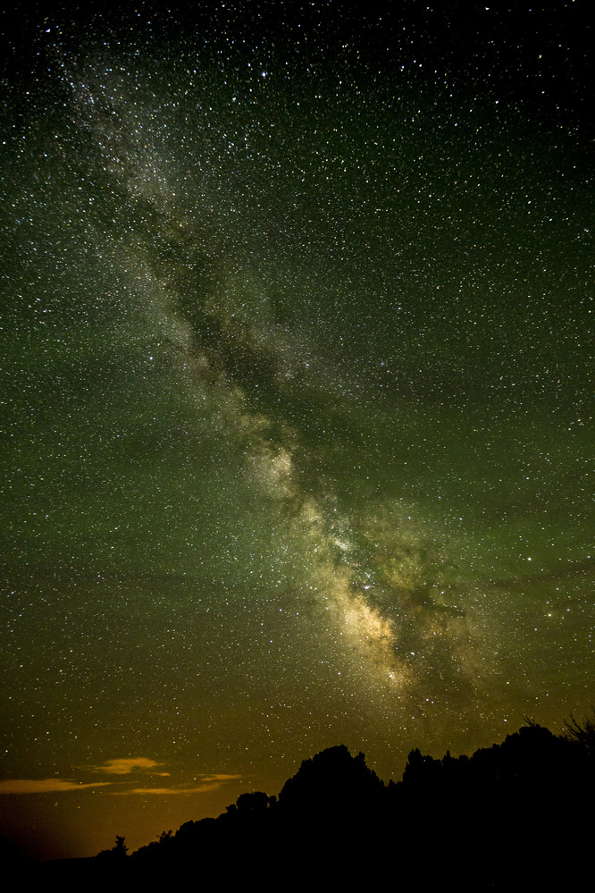 The Milky Way shines prevalent in the night sky from upper Left to lower right, with hills and cliffs silhouetted in the foreground.