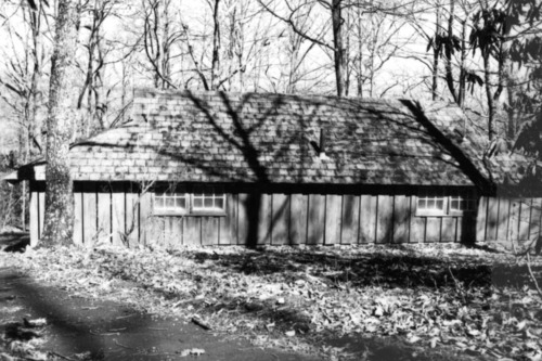 A single-story building in a wooded area has vertical wooden siding and a shingled roof. 