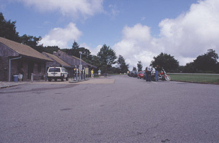 Vehicles and visitors collect along the front of a service building, with a white jeep by the gas pump.
