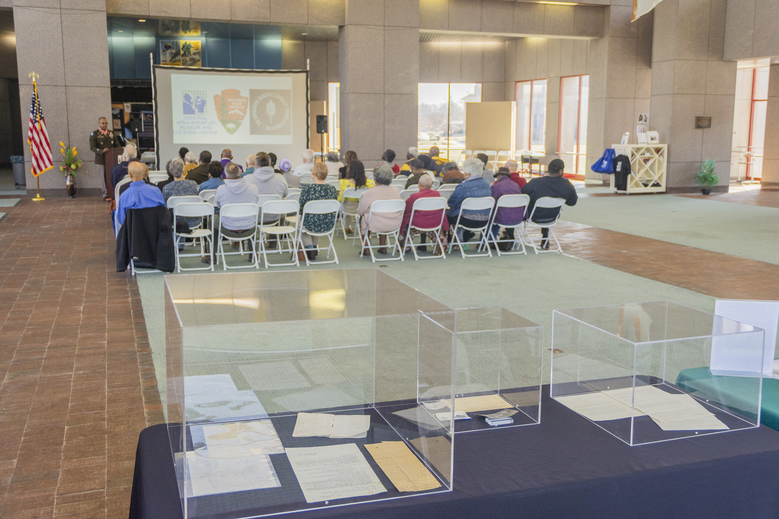 A man in a Army uniform standing behind a wooden podium speaking into a microphone atop the podium while seated guests in white chairs look and listen to him. Letters and documents lay on a blue-covered table with clear glass cubes cover and protect them.