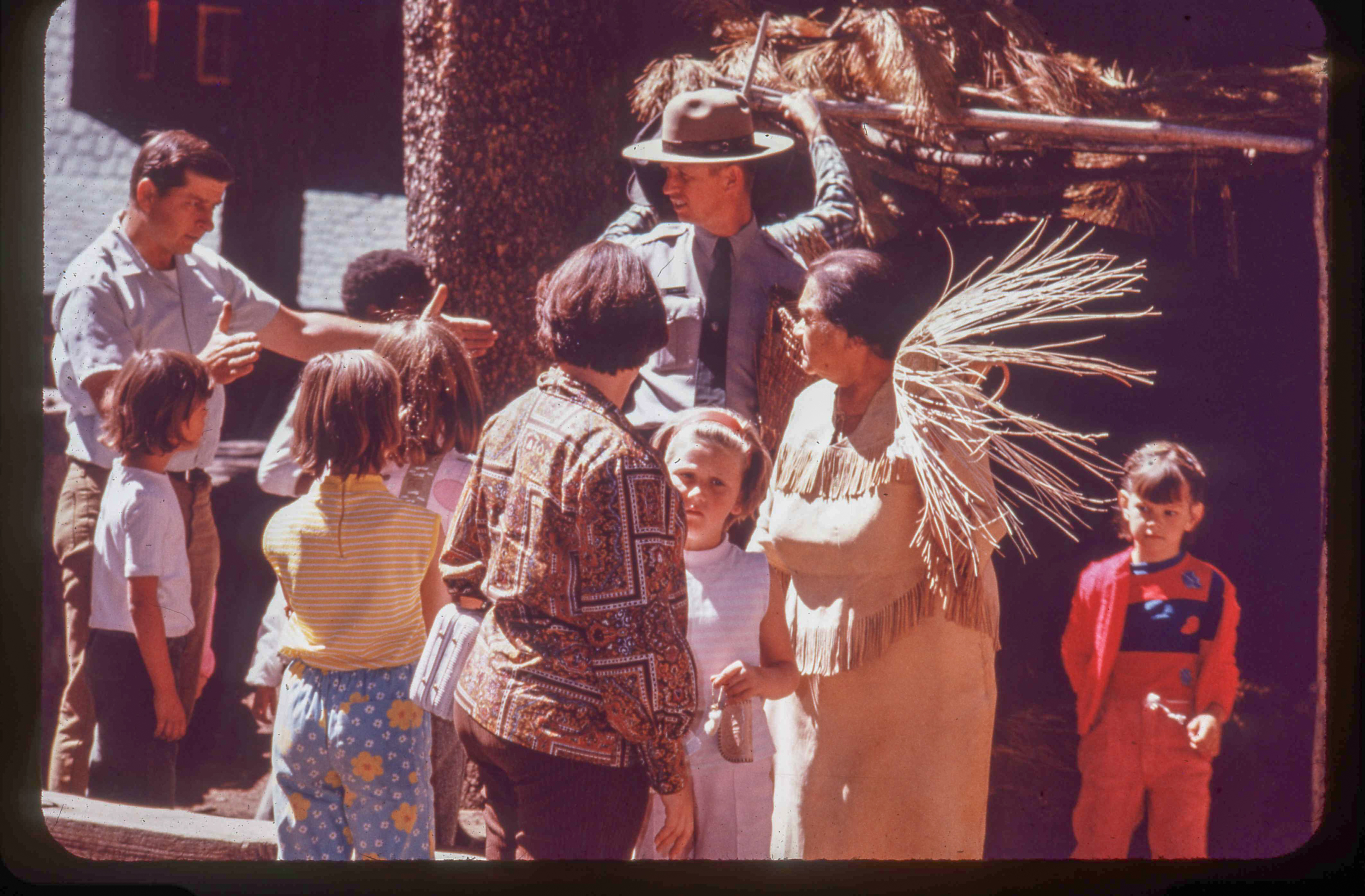 A woman in American Indian attire stands with a basket weaving in a demonstration to a group of visitors. A male park ranger stands to her right.