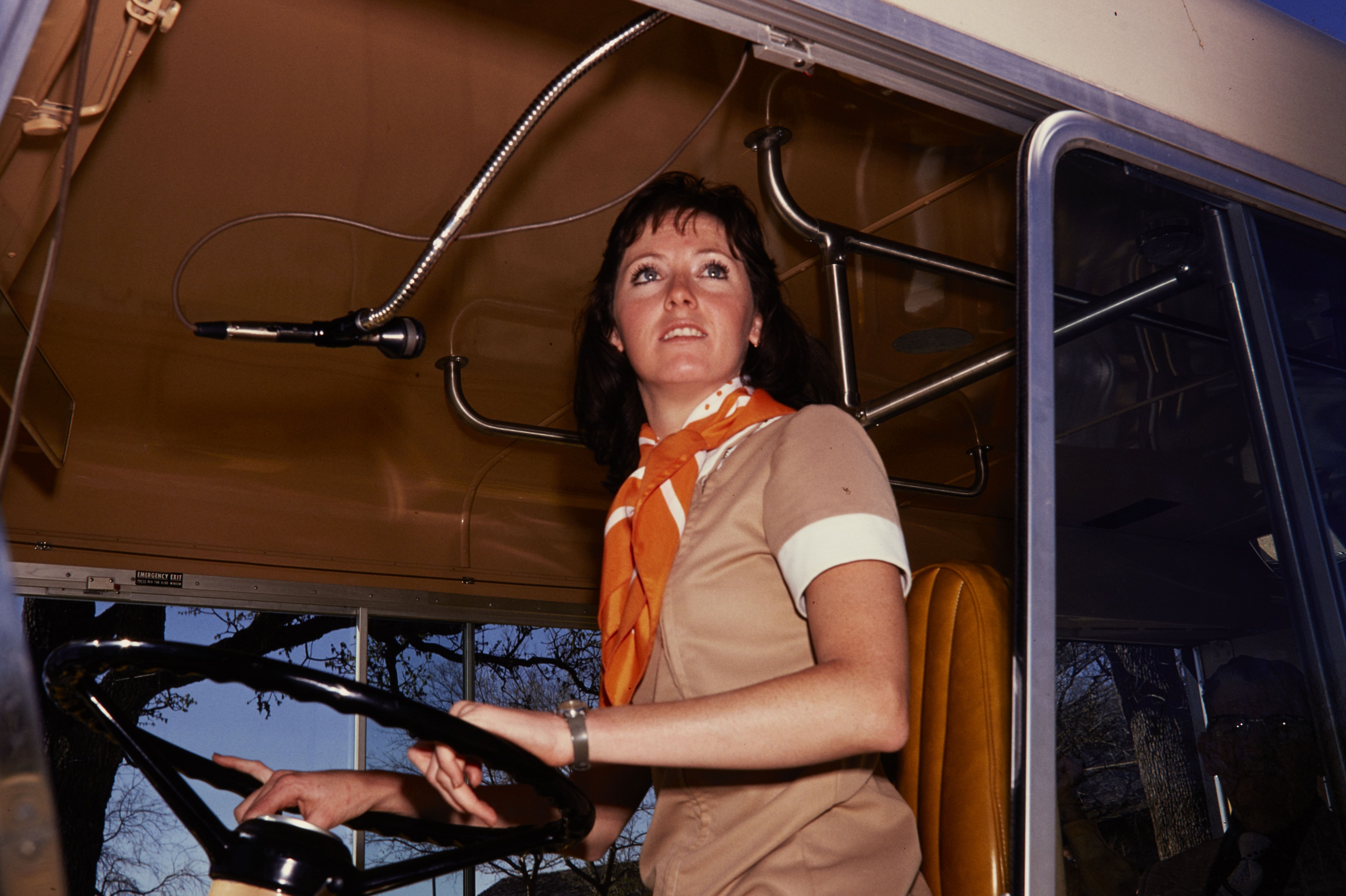 Woman in her NPS uniform sits in the driver's seat of a bus with her hands  on the steering wheel.