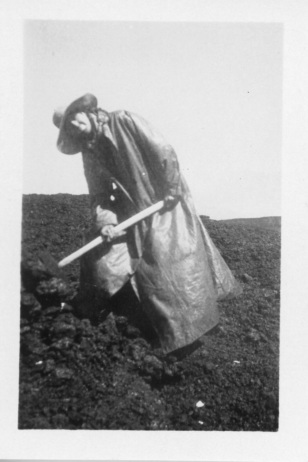 A black and white image of a man posing with a pickaxe on Mauna Loa. The man is located in the center of the image. He is wearing a cowboy hat, bandana, and a floor-length raincoat. He is leaning towards the left side of the image with his feet spread apart. The man is holding a pickaxe with both of his hands and is smiling at the camera. He is standing in a rocky, lava covered terrain.