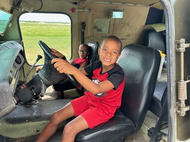 Two boys wearing black and red clothing sitting inside a green armored vehicle with a vast prairie in the background. 