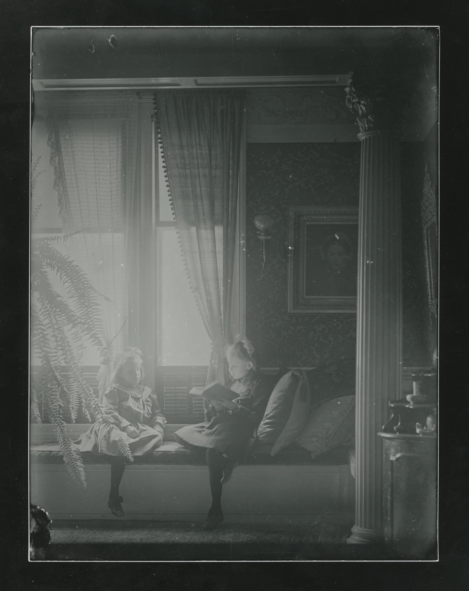 Two white girls sitting on a window seat bench in front of tall windows. One reads. Grecian column at right.