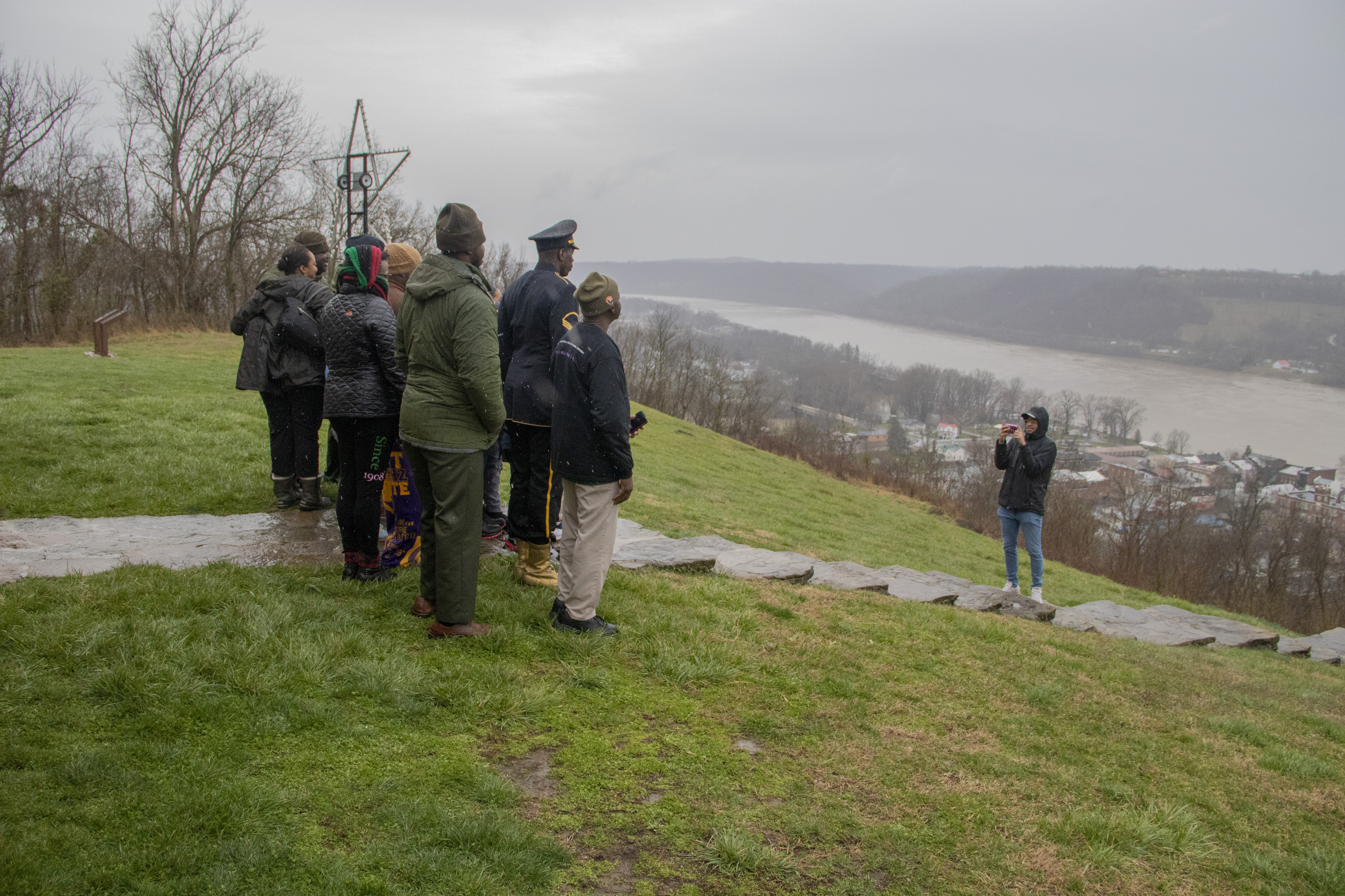 Several people standing on a sidewalk and grass for a photo with a long river in the background to the right.