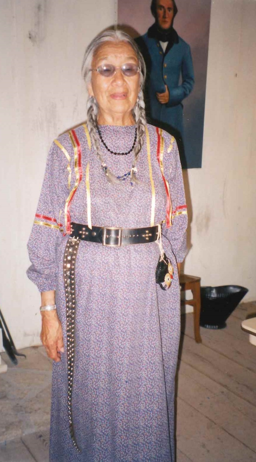 An American Indian woman with gray hair in braids wearing a purple dress with red and gold ribbon decoration