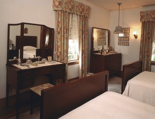 Bedroom looking over the footboards of two twin beds with two dressers and mirrors on opposite wall. Light blue walls and floral drapes.