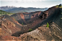 A large, steep-sided volcanic crater with stripes of black and reddish-brown around its sides and mountains in the distance.